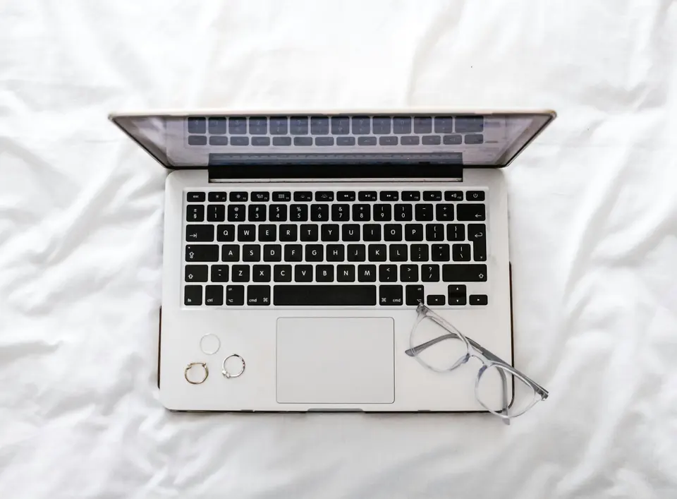 Laptop open on white sheets with glasses nearby