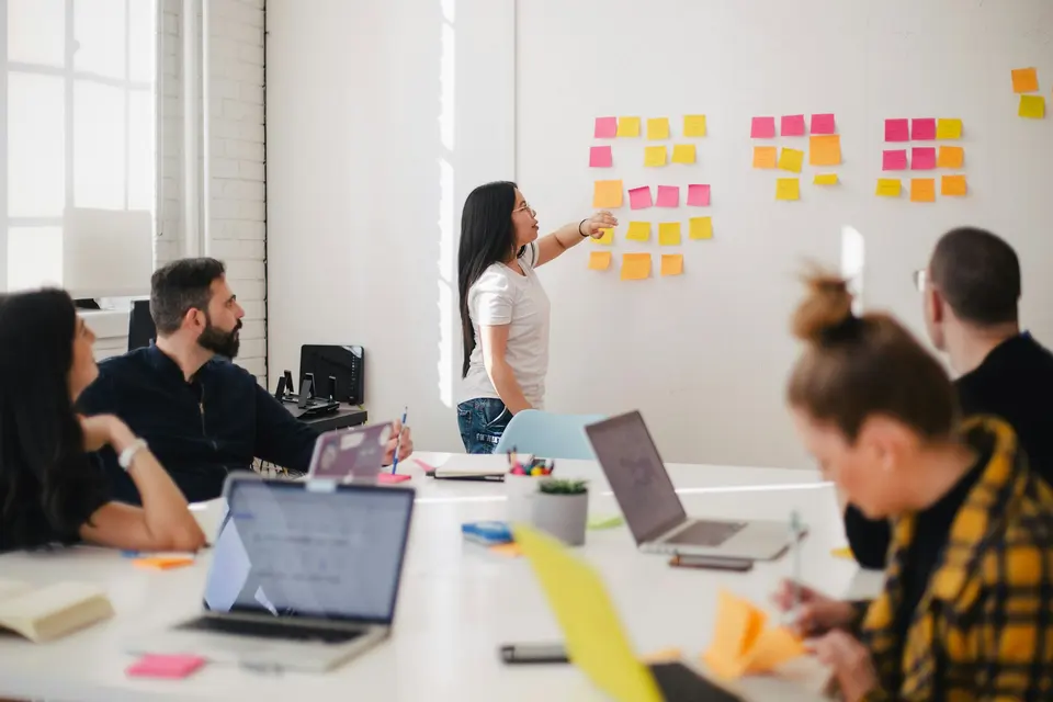 A group around a table while one person points to sticky notes on a wall
