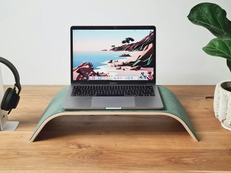 A laptop on a raised wooden stand beside headphones and a plant