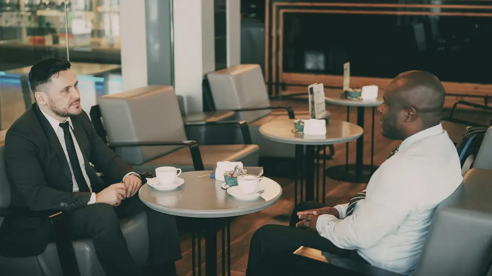 Two people seated across a cafe table with cups and a laptop