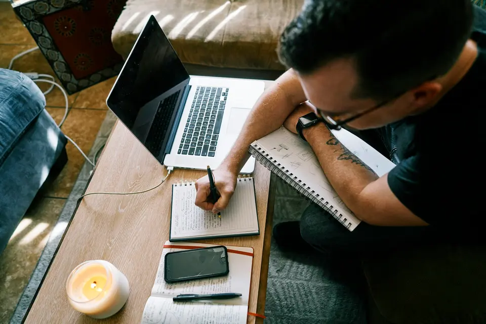 A person writing in a notebook beside a laptop and phone
